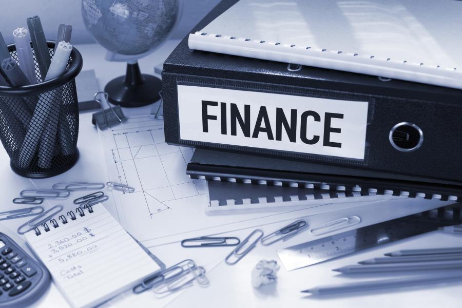 Photograph of a desk covered with a binder with "Finance" printed on the spine, notebooks, cup full of markers, globe, diagrams, paperclips, calculator, ruler, pencils, notepad with figures written on it