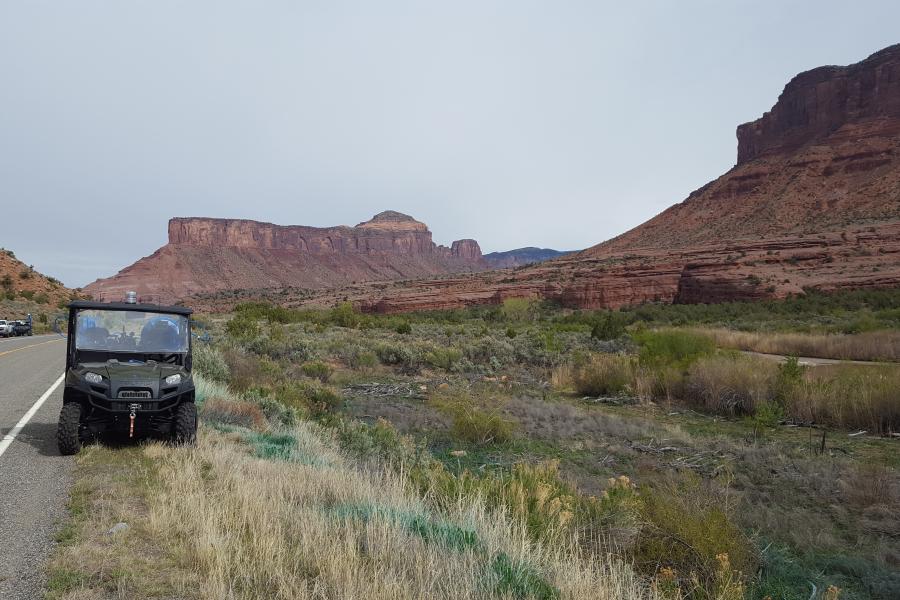 Photograph of Desert Landscape on the side of highway showing weed growth.  Jeep parked in front, mountain ranges in the background.