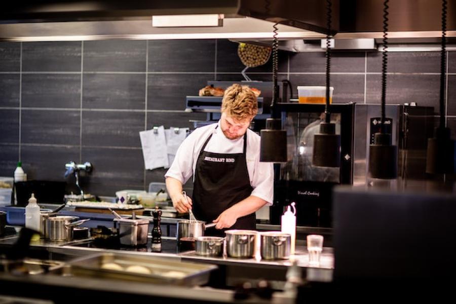 Chef cooking in kitchen with silver colored sauté pans over an island situated stove and in front of a black slate back wall