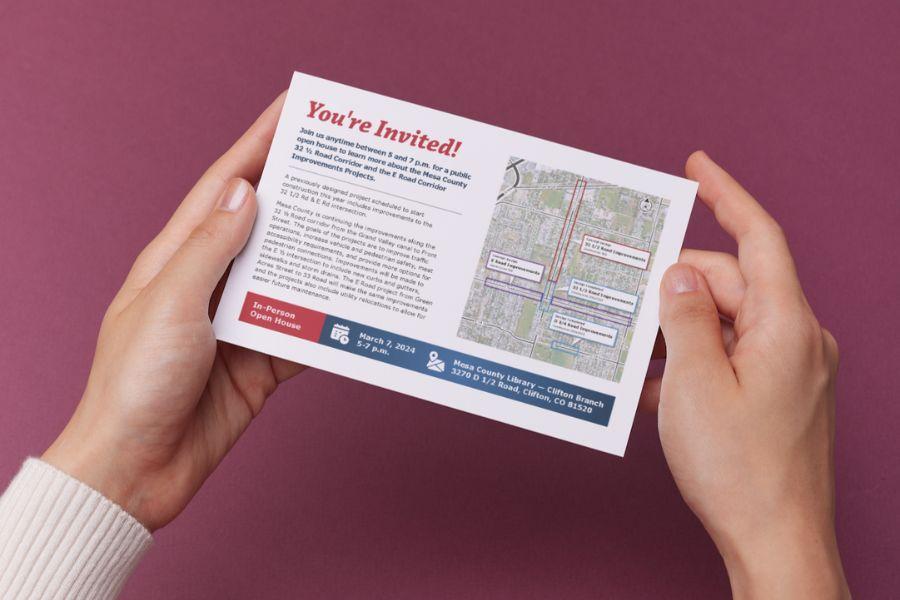 Hands holding white postcard displaying 32 1⁄2 Road Corridor and E Road Corridor Improvement Projects Open House details. 