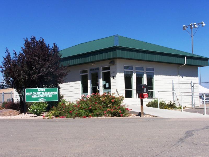 Photograph of the outside of Fairgrounds Office Building with Office Mesa County Fairgrounds / Mesa County Fair sign , mailbox with Daily Sentinel newspaper box, ramp to door with hand rail, flowering plants, and tree