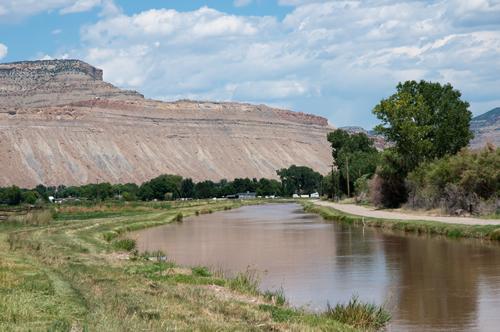 Photograph of Irigation ditch full of water with trees growing on the bank and the desert mountains and cliffs that make up the Bookcliffs that stretch almost 200 miles across western Colorado and eastern Utah.