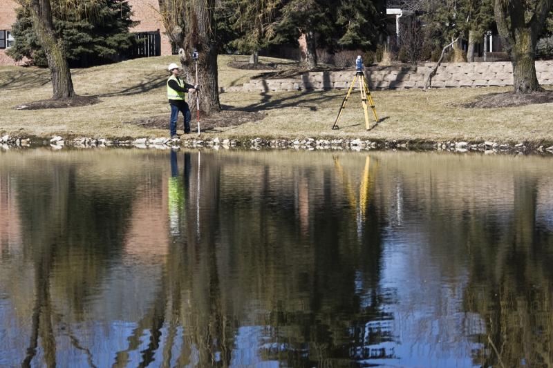 Photograph of Man with Survey Equipment by lake with building, grass, building in the background