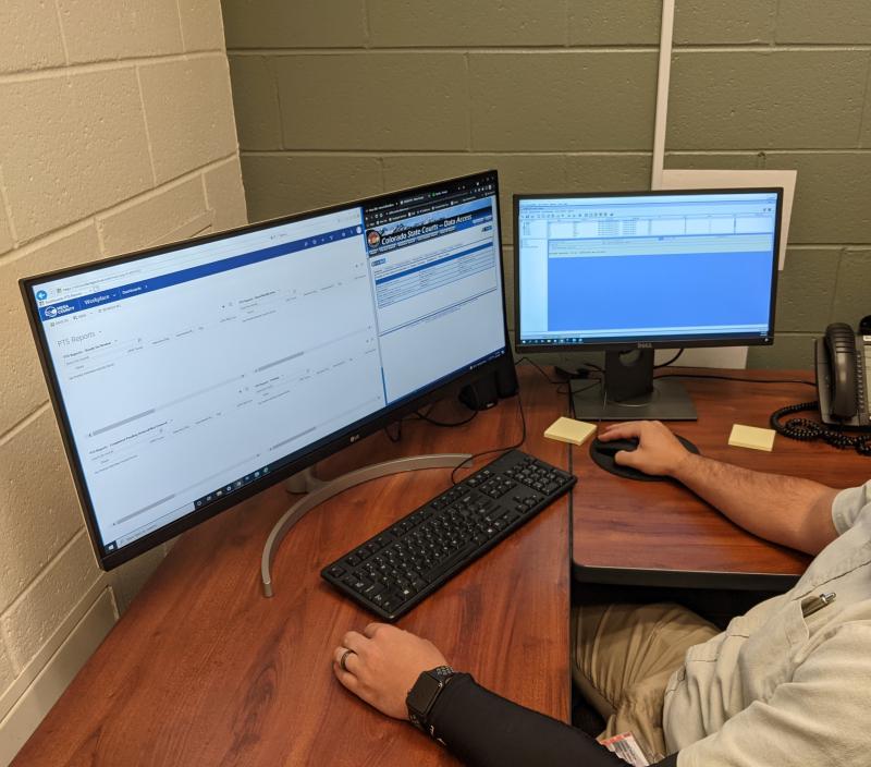 Photograph of a man sitting at a desk working on his computer.  With keyboard, mouse, two monitors, and telephone in the photograph.