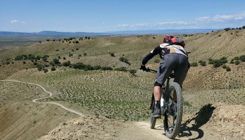 Rider going down knife edge trail, lots of desert and trail seen in the distance.