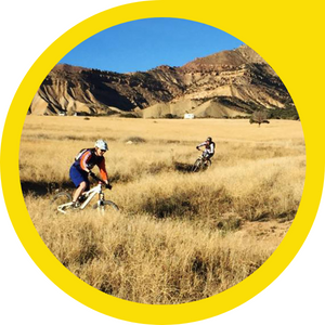 two mountain bikers weaving way through desert grasses in front of a small mountain range