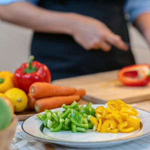 Chopping red, yellow, and green peppers  with other vegetables piled up next to the wood cutting board.