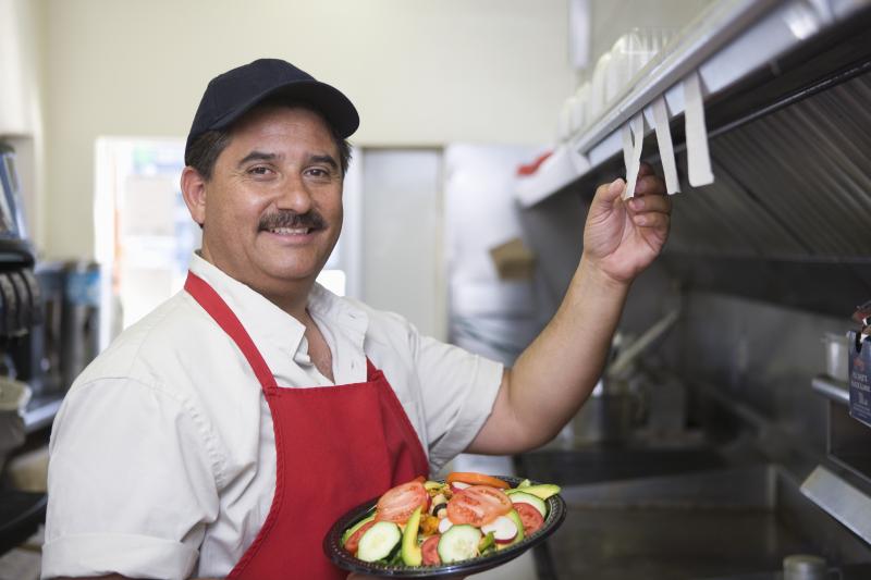 look cook with white shirt and red apron holding a plate of veggies and taking down ticket order