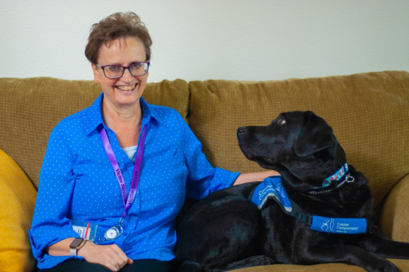 Rachael (black lab facility dog) and Ashley Edstrom (dog handler) sitting side by side on couch. 