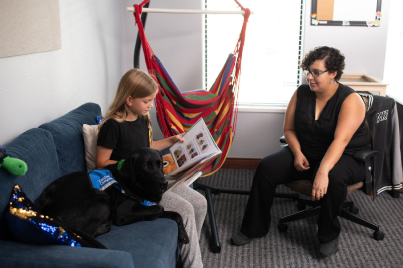 Rachael (black lab facility dog) sitting next to child while they read. 