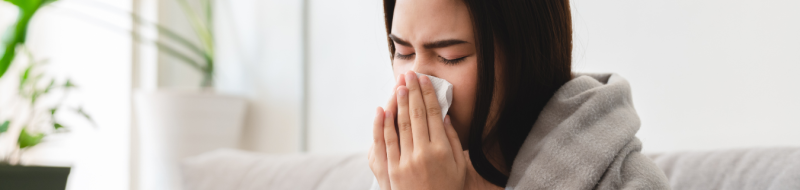 Woman on the couch with a blanket blowing her nose.