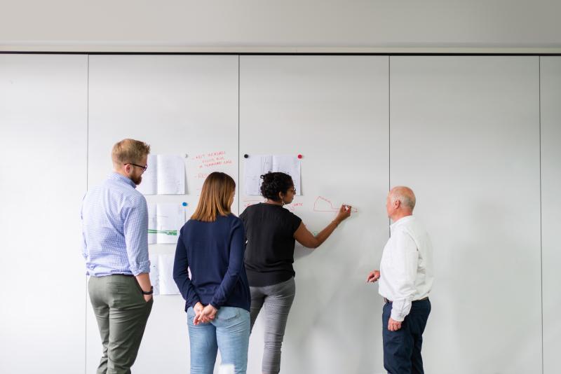 Four people stand in front of a large white board with their backs to the camera as a woman writes on the board in orange marker.