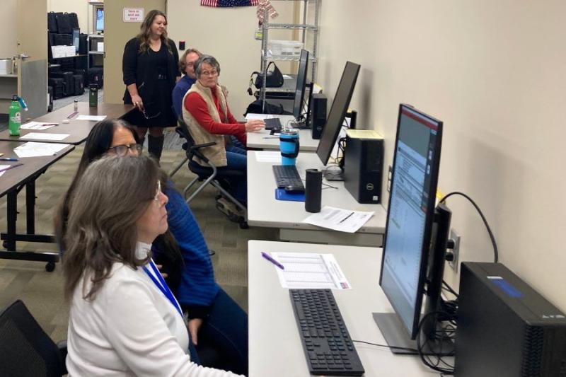 A row of people look at different computer screens as one woman walks behind them. 