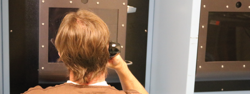 A male with light brown hair wearing a tan shirt is faced away from the camera. He is holding a phone to his ear and looking at the video visitation screen.