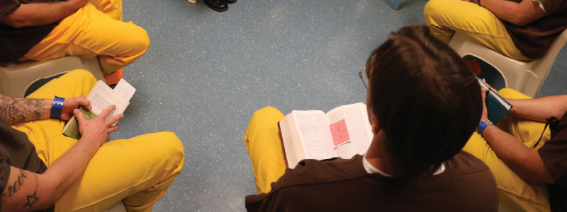 A group of male inmates sit in a semi-circle facing a male instructor during a Jail Based Behavioral Services program.