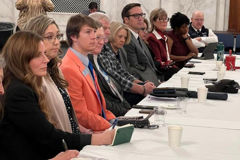 Several men and women sit at a white table and take notes. 