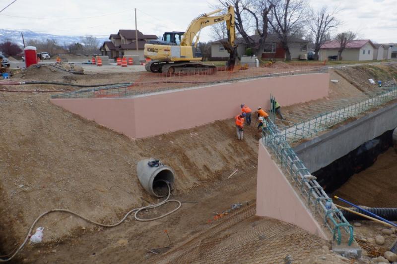 Five construction workers wearing bright orange and bright yellow/green stand in the dirt middle area of the unfinished bridge. 