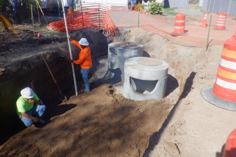 Two construction workers in yellow and orange clothing in a hole with orange construction cones around the site. 