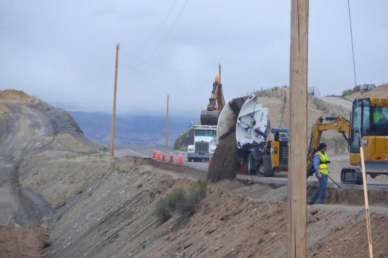 Construction worker on a hill working on the road beside it and a truck dumping dirt on the hill. 