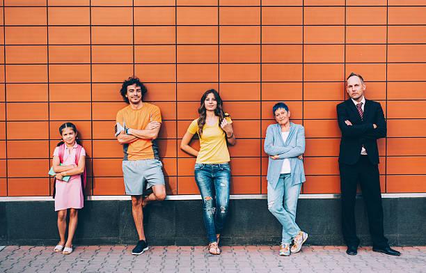 5 people of different ages leaning back against a brick wall. 