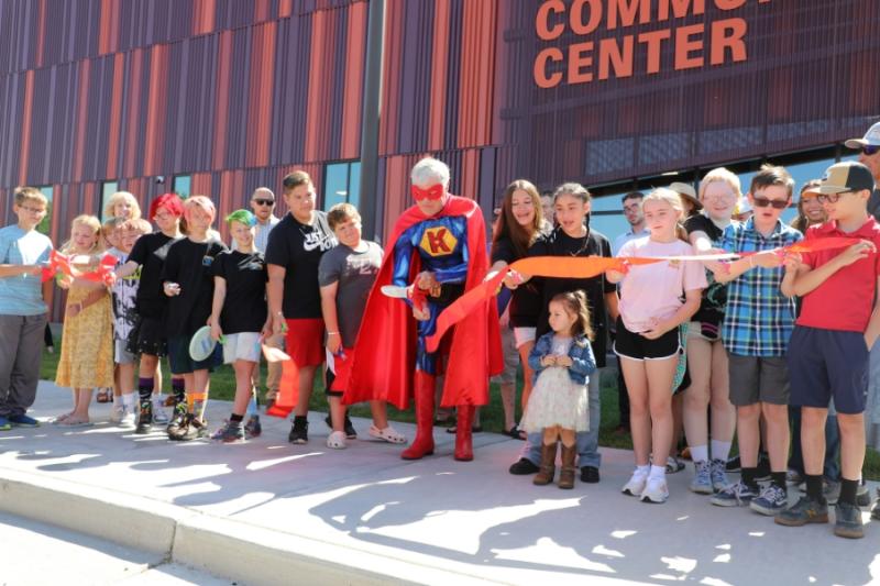 Large group of people standing around man dressed as superhero holding large red scissors. 