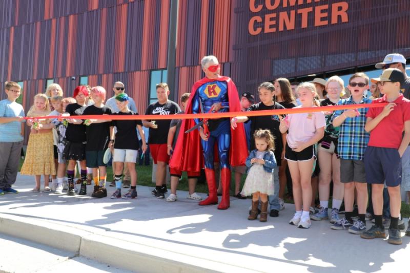 Large group of people standing around man dressed as superhero holding large red scissors. 