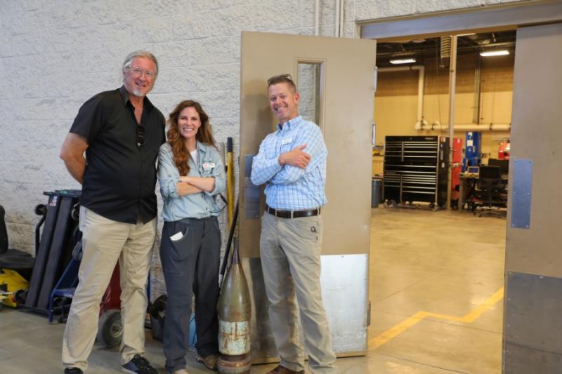 Three people standing next to very large bullet holding door open. 