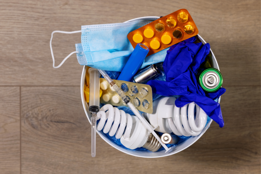 CFL lightbulbs, medication, batteries, syringes, and medical face mask sitting in trash can.