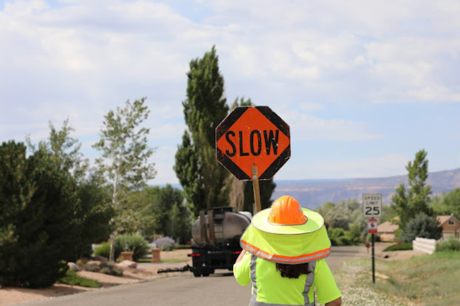 View from back of construction worker holding bright orange sign reading SLOW with trees and a truck in the background.