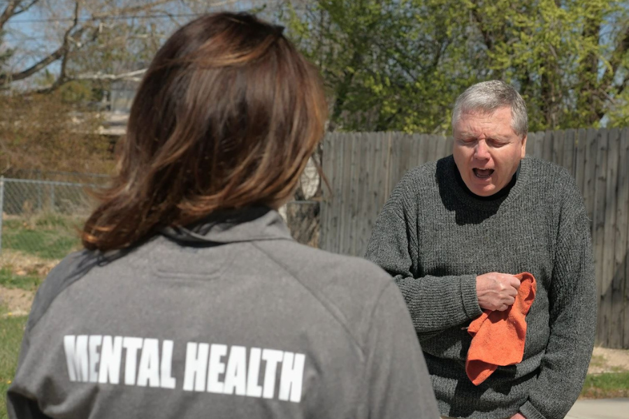 Back of woman's head and shirt reading "mental health" who is facing man with white hair in distress.