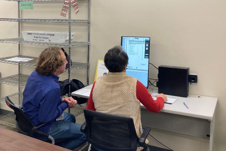 View from behind two people sitting in front of a computer. One man in a blue shirt and a woman in a red shirt with a tan vest. 