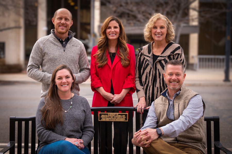 One man and two women stand behind a black bench as one woman and one man sit on the bench dedicated to a community member. 