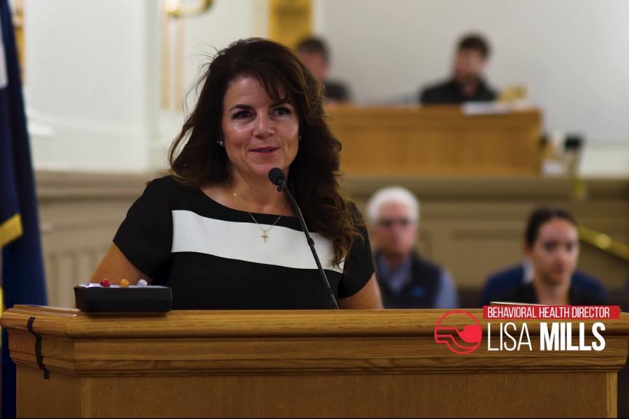 Woman stands at lectern with red and white text reading, "Behavioral Health Director Lisa Mills."