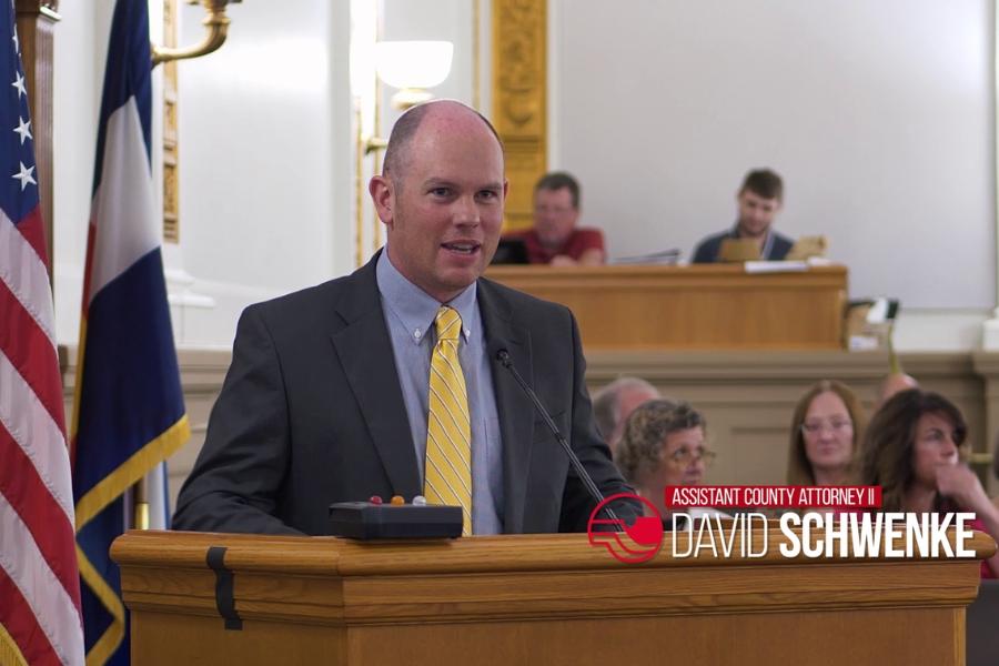 Man stands at lectern with red and white lower third reading, "Assistant County Attorney 2 David Schwenke."