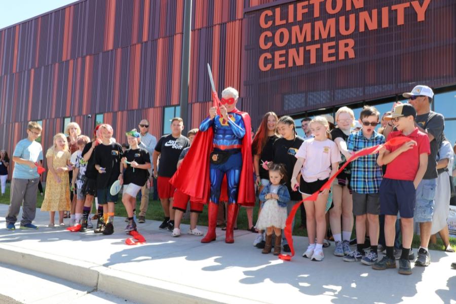 Large group of people standing around man dressed as superhero holding large red scissors. 